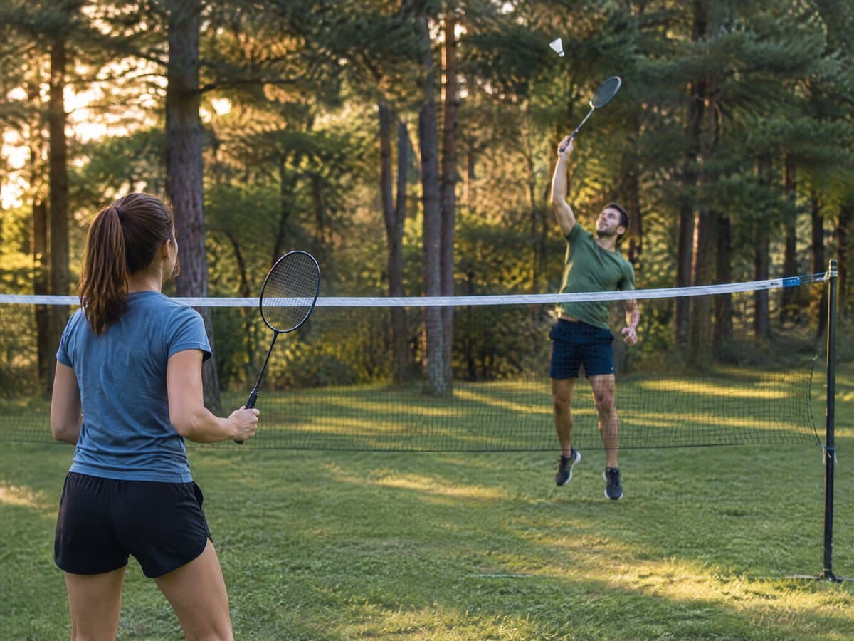 A woman and a man playing badminton over a net among tall pine trees, the man jumping to hit a white shuttlecock
